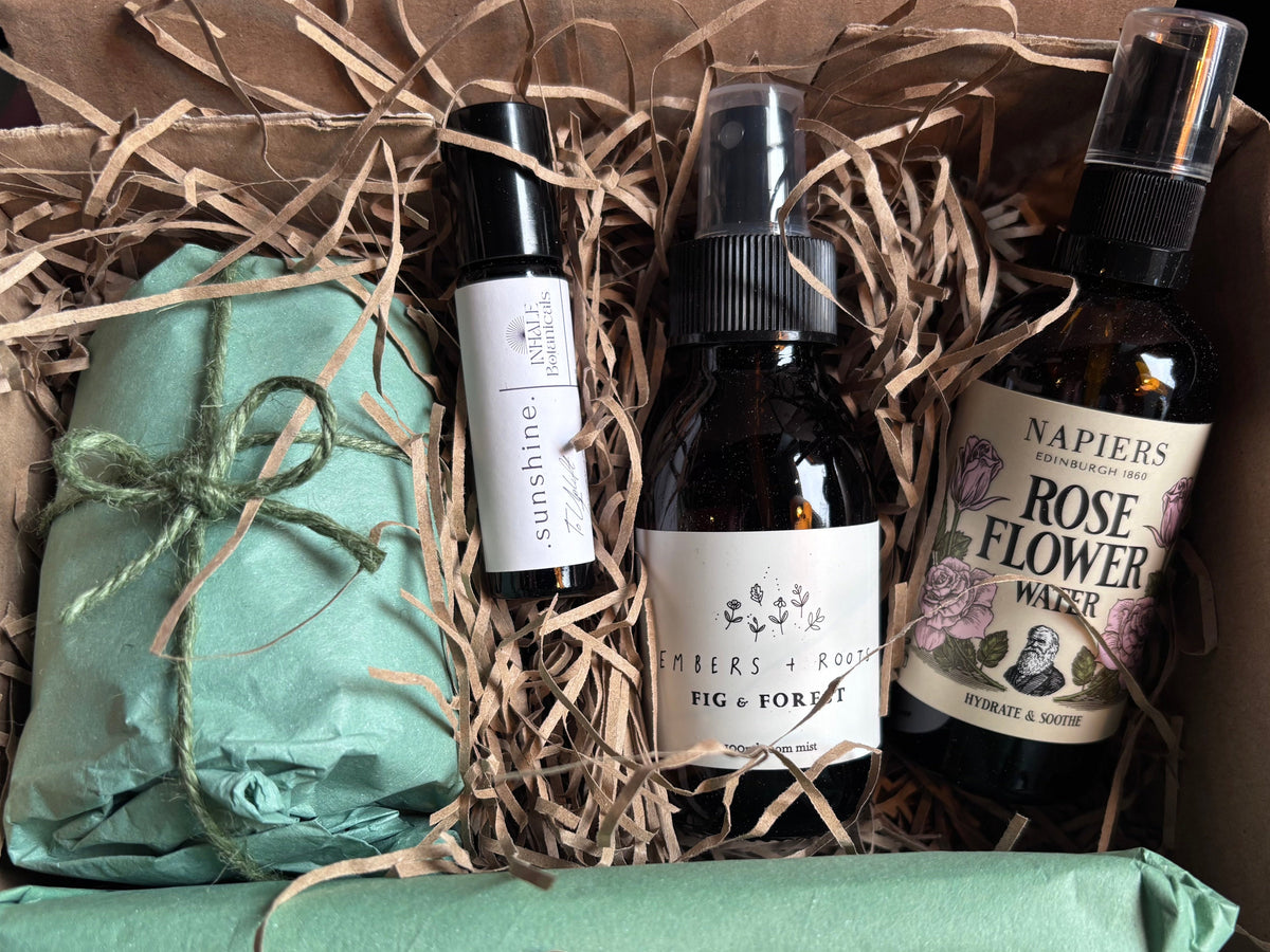 A Holme Botanicals gift box set containing two sage green wrapped items tied with green twine, a small aromatherapy roll on bottle labeled 'sunshine', a dark amber room spray labeled 'fig & forest' from Embers & Roots, and a dark amber bottle of Face Spritz labeled 'NAPIERS ROSE OF THE FLOWER WATER', all arranged in brown paper shred inside a cardboard box.