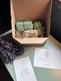 An open cardboard luxury ritual gift box containing Holme Botanicals products: wrapped bars of soap in green and beige paper wrapping, placed on a pale green tabletop next to two branded cards and a sprig of dried lavender. The cards show the brand Holme Botanicals.